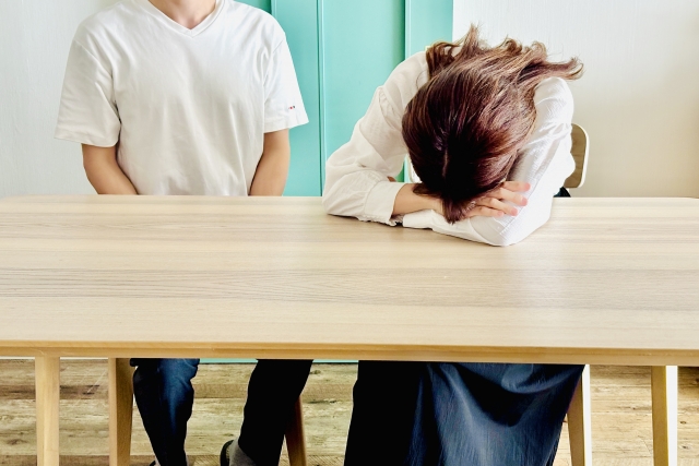 机に顔を伏せる女性とそれを見ている男性