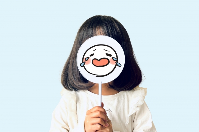 泣いている顔文字の札を自分の顔に当てる女の子