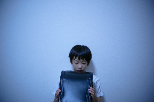 暗い部屋でランドセルを抱え込み下を見ている男の子